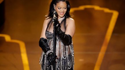Rihanna, dressed in a silver and black dress, stands behind a microphone, smiling.