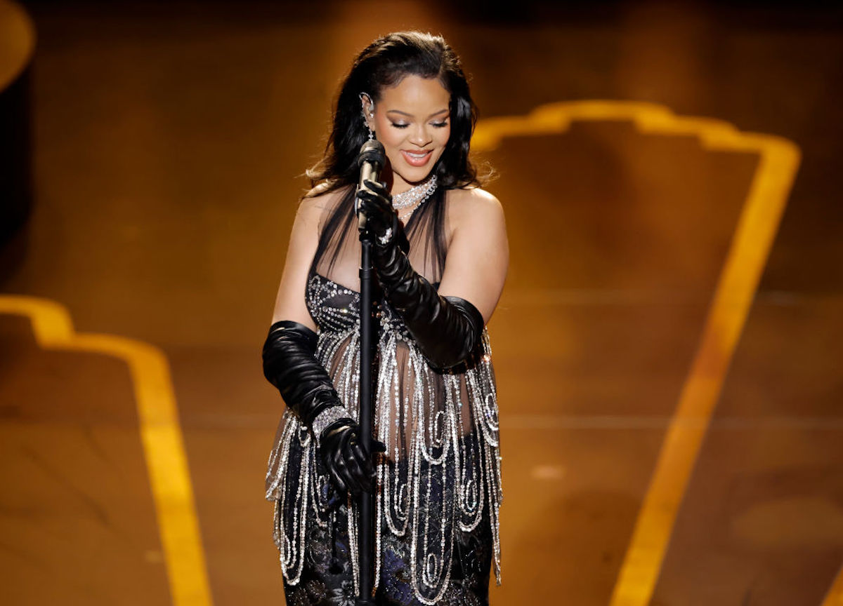 Rihanna, dressed in a silver and black dress, stands behind a microphone, smiling.