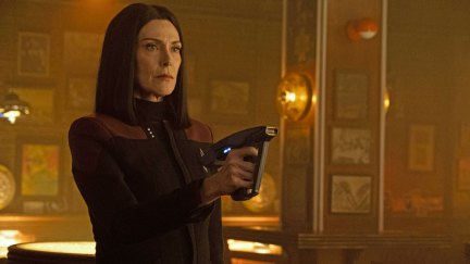 Image of Michelle Forbes as Ro Laren on 'Star Trek: Picard.' She is standing in a bar pointing a phaser at an unseen person. Her black, shoulder-length hair is parted down the middle, and she's wearing a black Starfleet uniform with dark red on the shoulders and three pips (Commander).