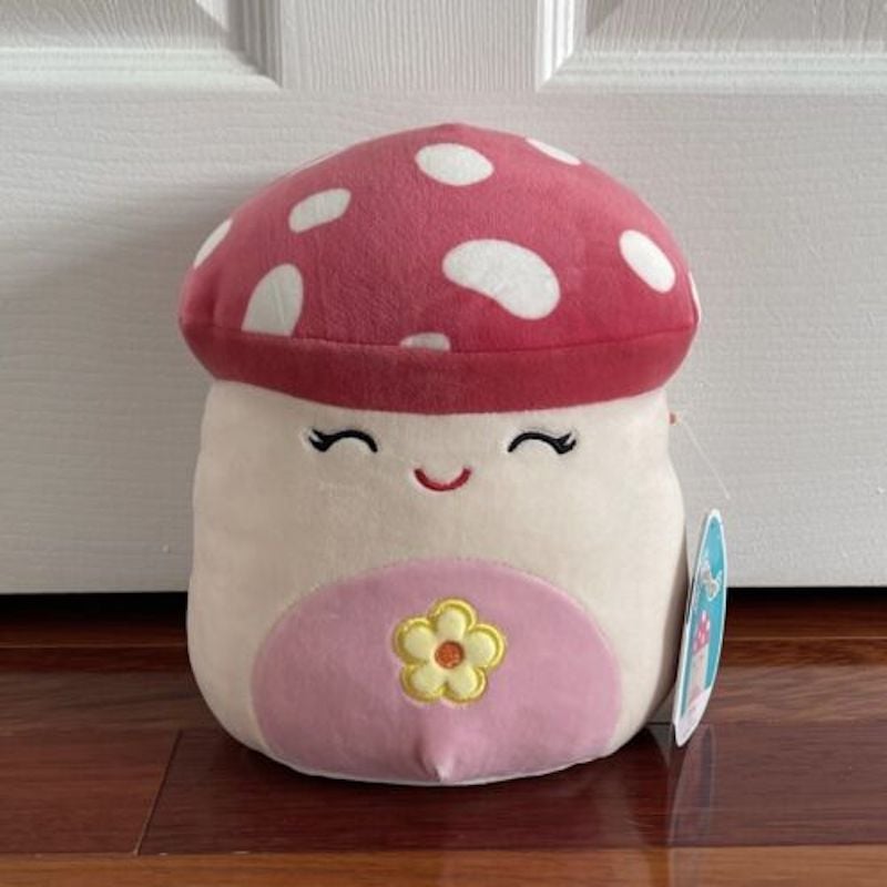 A mushroom squishmallow with a white spotted red cap, and a pink stomach with a yellow flower on it