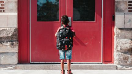 A young child stands outside closed school doors.
