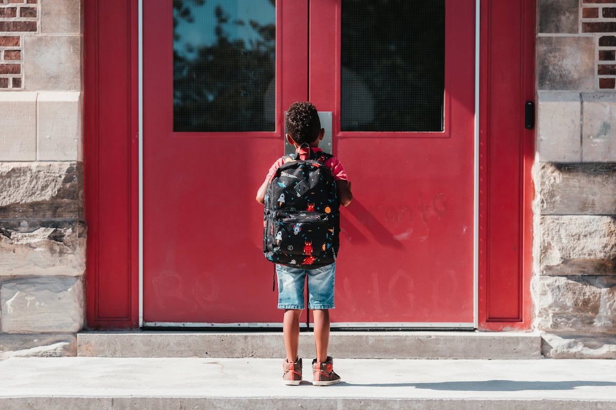 A young child stands outside closed school doors.