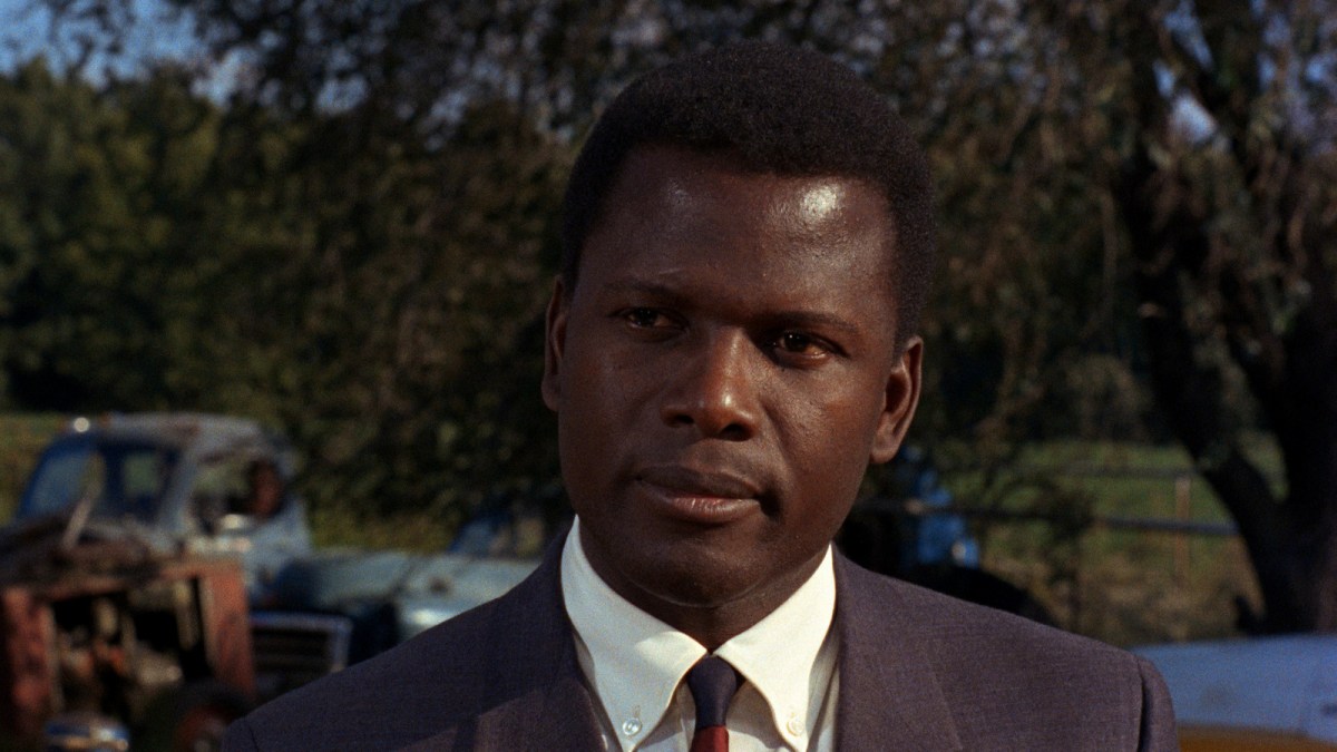 Image of Sidney Poitier as Virgil Tibbs in 'In the Heat of the Night.' We see him from the shoulders up. He's standing outdoors in front of trees and a farm truck. He's wearing a brown suit with a white collared shirt and a tie. He has short, black hair. He's looking at something suspiciously. 