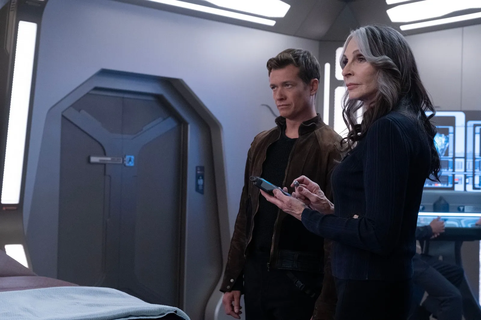Ed Speleers as Jack Crusher and Gates McFadden as Beverly Crusher on 'Star Trek: Picard.' They are standing close to each other to the left of a bed in sickbay. They are both looking at something in the distance. Beverly is in the foreground  holding a tricorder. She's dressed all in navy blue and has long salt-and-pepper hair. Jack stands to her right. He has short, brown hair, and is wearing a brown jacket over a black shirt. 