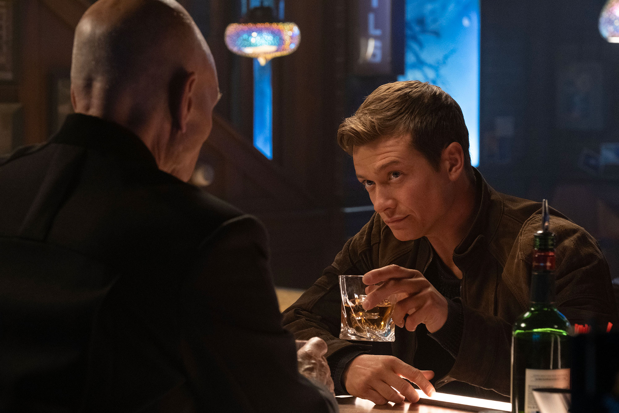Image of Ed Speleers as Jack Crusher on 'Star Trek: Picard.' He is standing opposite Patrick Stewart as Jean-Luc Picard, who has his back to the camera. Jack is sitting at a bar holding up a glass of whiskey. He has short, light brown hair and is wearing a brown jacket. Jean-Luc is bald and wearing all black. 