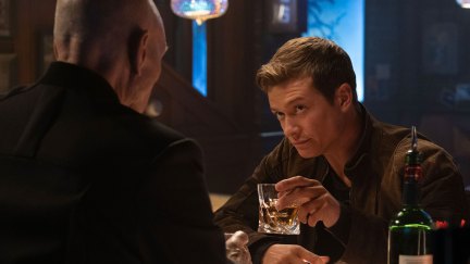 Image of Ed Speleers as Jack Crusher on 'Star Trek: Picard.' He is standing opposite Patrick Stewart as Jean-Luc Picard, who has his back to the camera. Jack is sitting at a bar holding up a glass of whiskey. He has short, light brown hair and is wearing a brown jacket. Jean-Luc is bald and wearing all black.