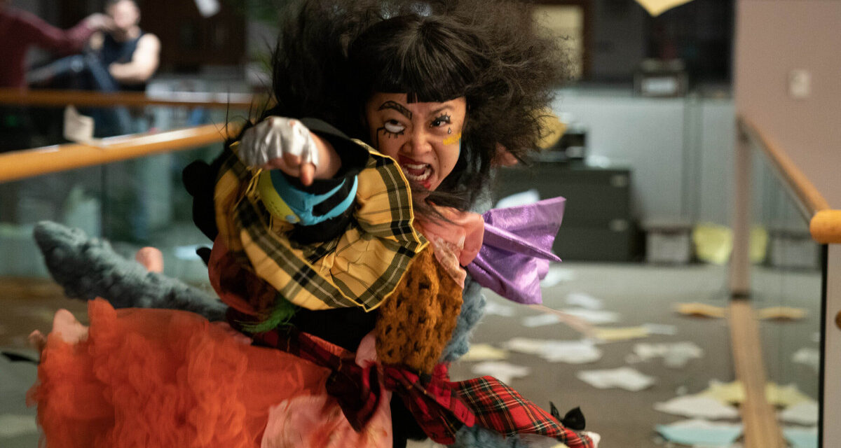Image of Stephanie Hsu as Jobu Tupaki in 'Everything, Everywhere, All at Once." She is wildly dressed in an enormous, colorful, patchwork dress as she throws a punch while flying through the air. Her dark hair is a huge mess of hair orbiting her head. She's in an office, and papers are scattered all over the floor. 
