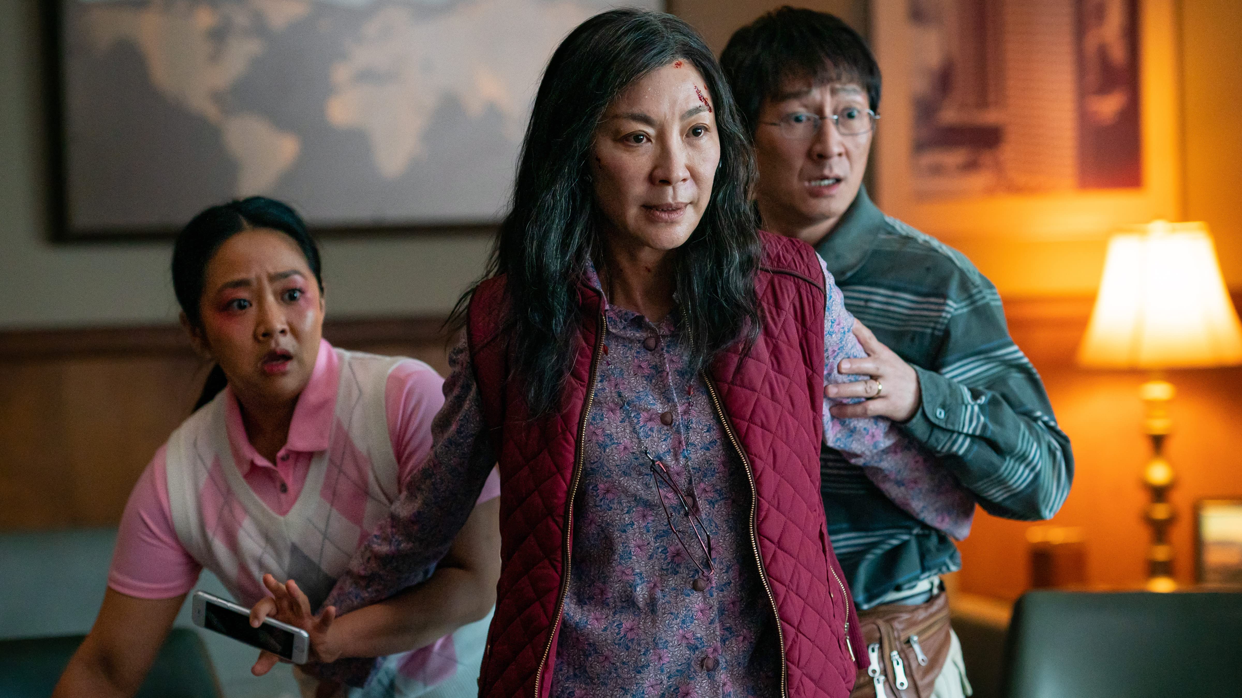 Image of Stephanie Hsu (Joy), Michelle Yeoh (Evelyn), and Ke Huy Quan (Waymond) in 'Everything, Everywhere, All at Once.' Evelyn is in the foreground with her arms out on either side to protect Joy and Waymond. Evelyn has long, black hair, and is wearing a maroon quilted vest over a purplish flower-print shirt. Joy cowers behind her on the right, holding her phone. Her long, dark hair is in a ponytail, and she's wearing a pink polo shirt under a pink, plaid pullover. Waymond is on Evelyn's right wearing a striped, green button-down and a brown fanny pack. He's wearing glasses and his dark hair is short.