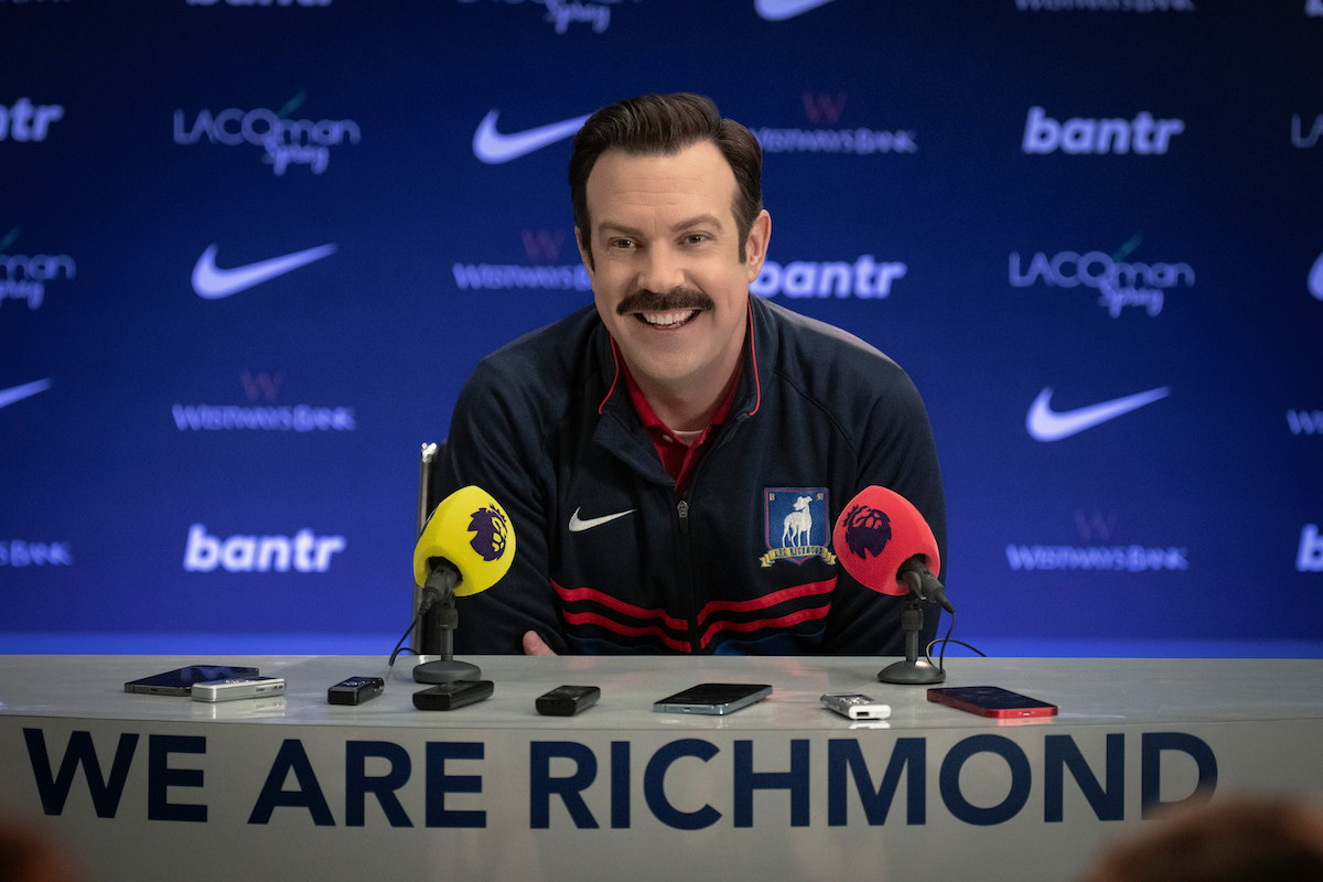 Ted Lasso smiles behind a podium at a press conference. The podium says "We Are Richmond."