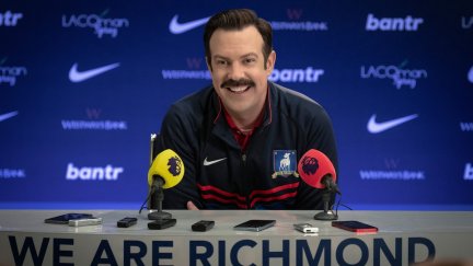 Ted Lasso smiles behind a podium at a press conference. The podium says 