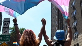 Two people hold hands raised in the air while waving a blue, pink, and white trans flag above them