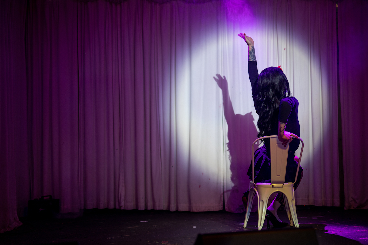 A drag performer sits in a chair on a stage with her arm in the air, lit by a spotlight.