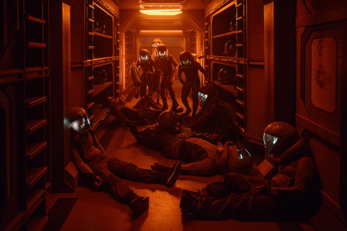 Image from the SyFy show, 'The Ark.' A spaceship corridor bathed in red light. In the foreground are several passengers passed out in space suits. Approaching them are 4 passengers in space suits trying to get past the bodies.