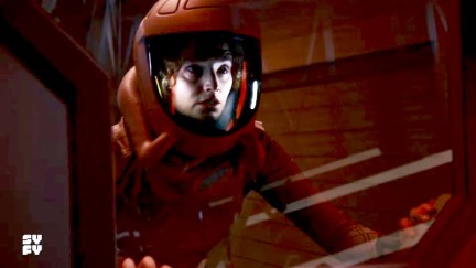 Image of Christie Burke as Sharon Garnet on the SyFy show 'The Ark.' She is a white woman in a red space suit looking out the window of a large spaceship. She looks worried.