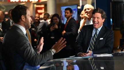 Tucker Carlson cackles from a news desk during a live taping, surrounded by a crowd of onlookers.