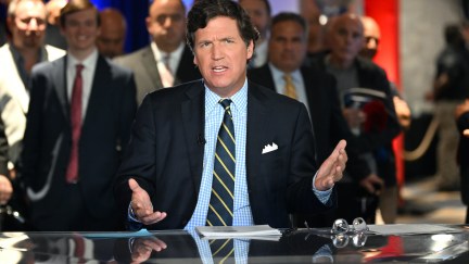 Tucker Carlson sits at a news desk, with a crowd of men in suits in the background watching him.