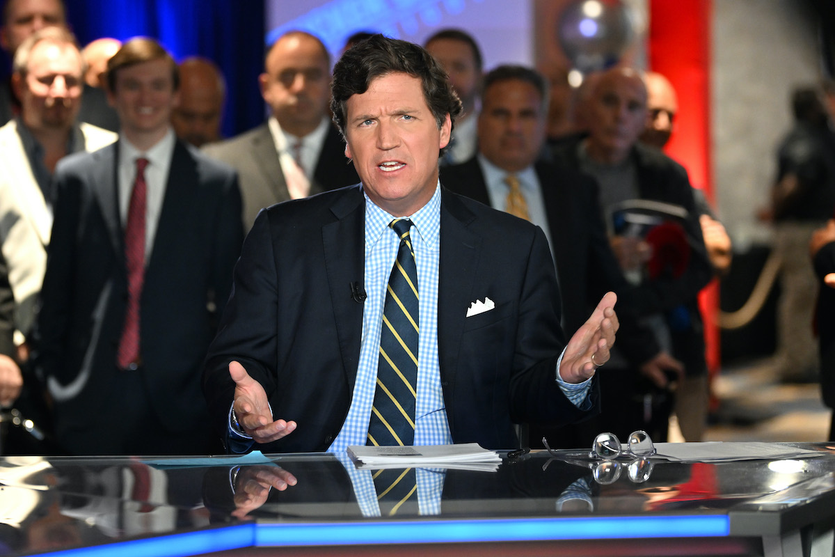 Tucker Carlson sits at a news desk, with a crowd of men in suits in the background watching him.