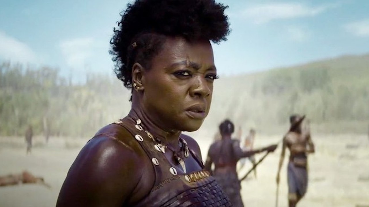 Viola Davis in a scene from 'The Woman King.' We see her from the shoulders up. She's standing outdoors and looking into the distance. Her hair is shorter on the sides and puffed high down the middle of her head like a mohawk. She has a scar over her eyebrow, and is wearing a sleeveless, brown, leather military uniform and a bone necklace.
