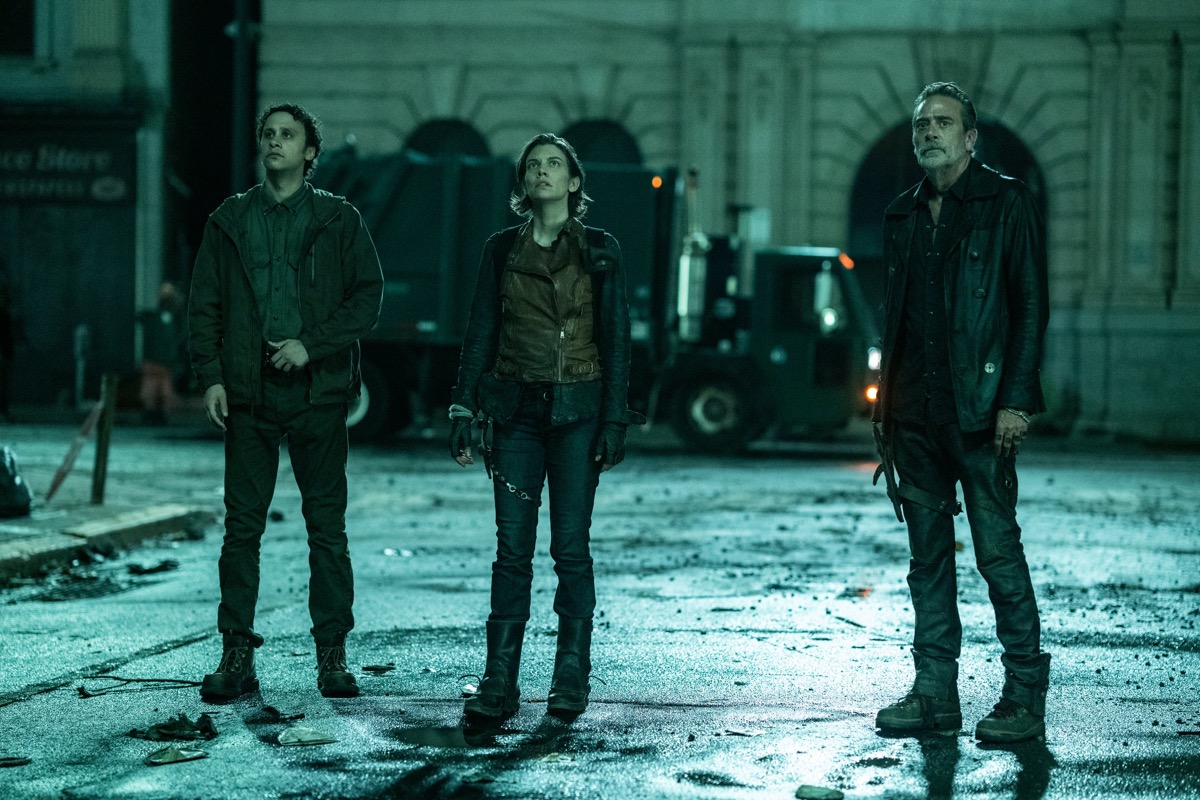 Trey Santiago-Hudson as Jano, Lauren Cohan as Maggie Rhee, Jeffrey Dean Morgan as Negan in a scene from 'The Walking Dead: Dead City' on AMC. This is a full-body image, and they are standing in the middle of a New York street at night with a truck in the background. They're all looking up at something. Jano (left) has brown skin and curly, dark hair. He's wearing a dark button-down-, a brownish-greenish jacket, and dark pants and boots. Maggie (middle) is a white woman with chin-length, dark hair wearing a black, long-sleeved shirt, black pants and boots, and a brown leather vest. Negan (right) is a white man with slicked-back dark hair and a salt-and-pepper beard. He's wearing all black, topped off with a black leather jacket.