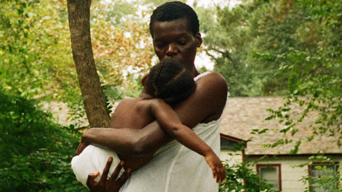 A Black woman cradles a child while standing in front of a house surrounded by lush greenery in 'All Dirt Roads Taste of Salt'