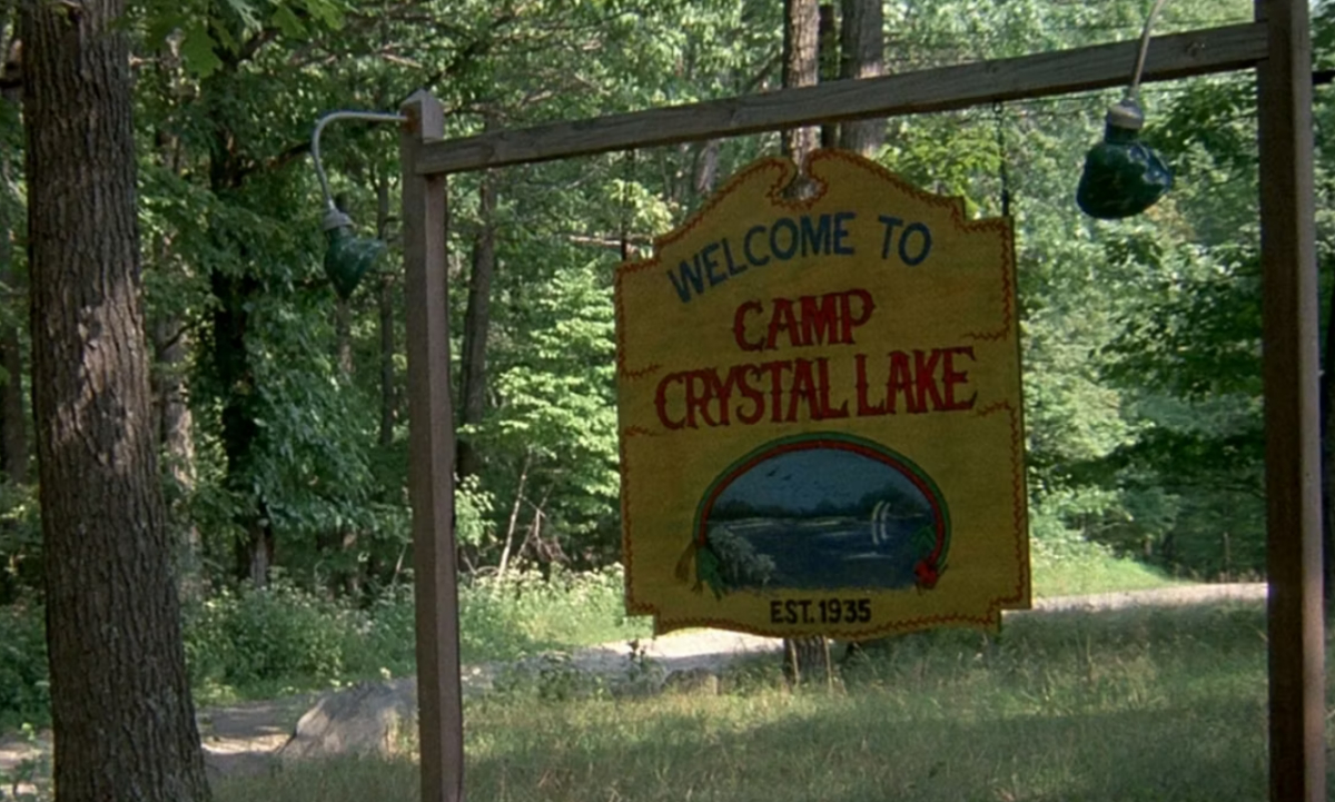 A sign hanging from a post in the woods reads "Welcome to Camp Crystal Lake - Est. 1935" in 'Friday the 13th'
