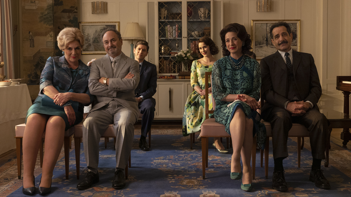 (left-right) Caroline Aaron as Shirley Maisel, Kevin Pollak as Moishe Maisel, Michael Zegan as Joel Maisel, Rachel Brosnahan as Midge Maisel, Marin Hinkle as Rose Weissman and Tony Shaloub as Abe Weissman in a scene from Amazon's 'The Marvelous Mrs. Maisel.' They are all seated in chairs at an indoor reception. The parents are seated in the front row, each couple on opposite sides of an aisle. Joel and Midge are seated one row behind them on opposite sides of the aisle. 