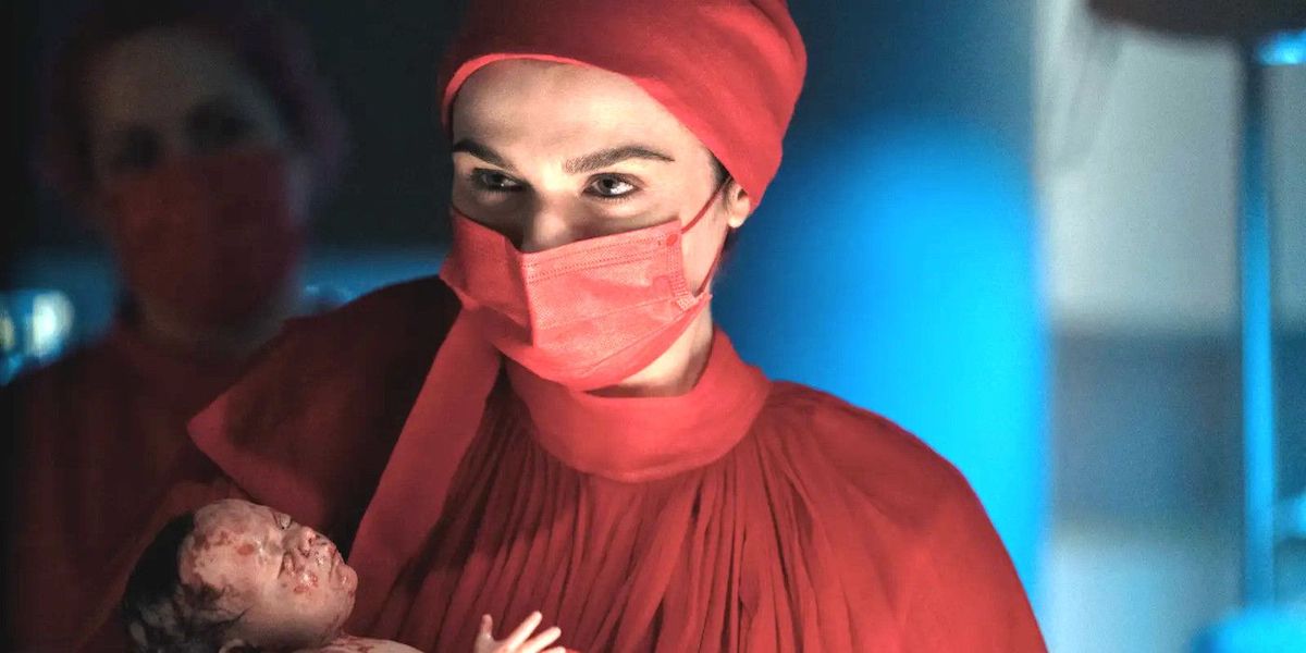 Rachel Weisz in a red gown and mask, holding a newborn baby.