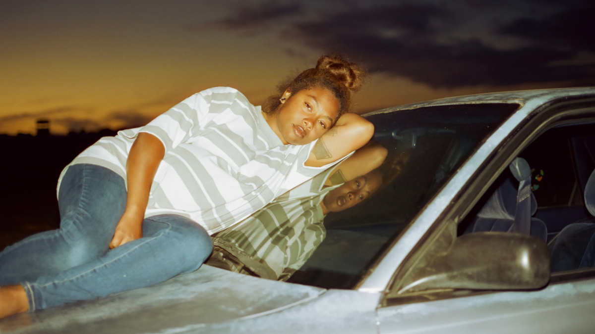 In 'Earth Mama,' a woman (Tia Nomore) reclines on the hood of a white car at sunset. She has a tired expression on her face.