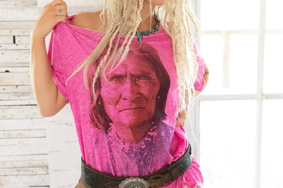A blonde woman with lots of thin, messy braids wear a pink t-shirt with a black and white picture of Goyaałé on it.