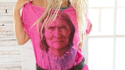 A blonde woman with lots of thin, messy braids wear a pink t-shirt with a black and white picture of Goyaałé on it.