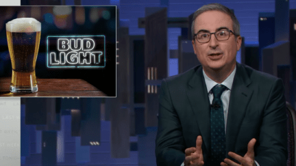 John Oliver sits next to a sign for Bud Light