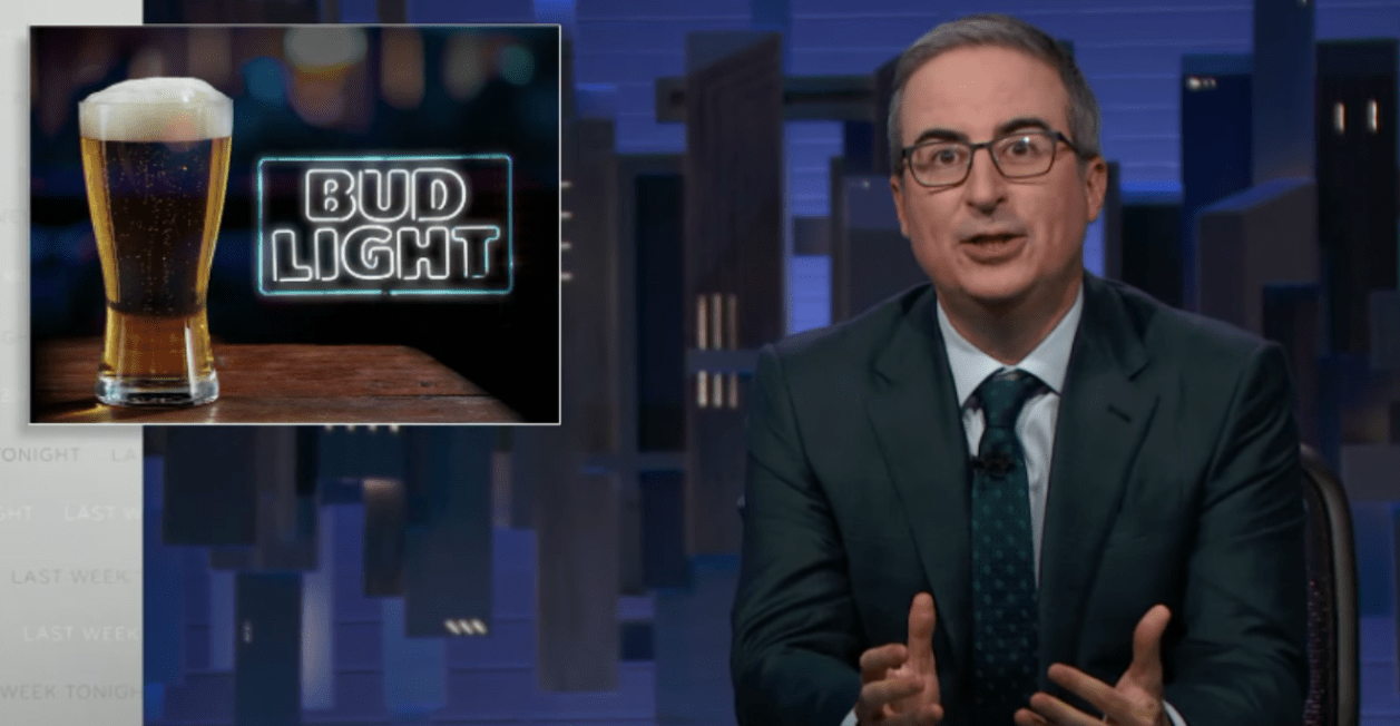 John Oliver sits next to a sign for Bud Light