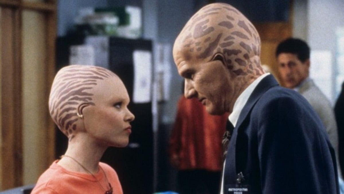 Lauren Woodland as Emily and Eric Pierpoint as George on FOX's 'Alien Nation.' They are both "Newcomer" aliens, and so both are bald with spotted heads. They are both portrayed by white actors and standing in profile facing each other. Emily is a teenage girl wearing a necklace and an orange shirt. George is an adult man wearing a navy blue suit with a white buttondown. They are in an office.