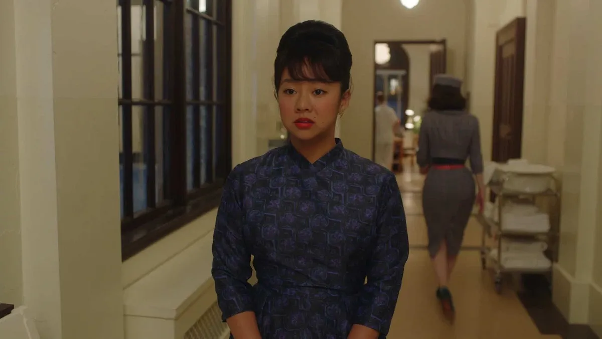 Stephanie Hsu as Mei in a scene from Amazon's 'The Marvelous Mrs. Maisel.' She's a young, Chinese woman with her black hair back in a bun and bangs across her forehead. She's wearing a patterned, purple dress with a high neckline. She looks concerned as she walks down a hospital hallway. 