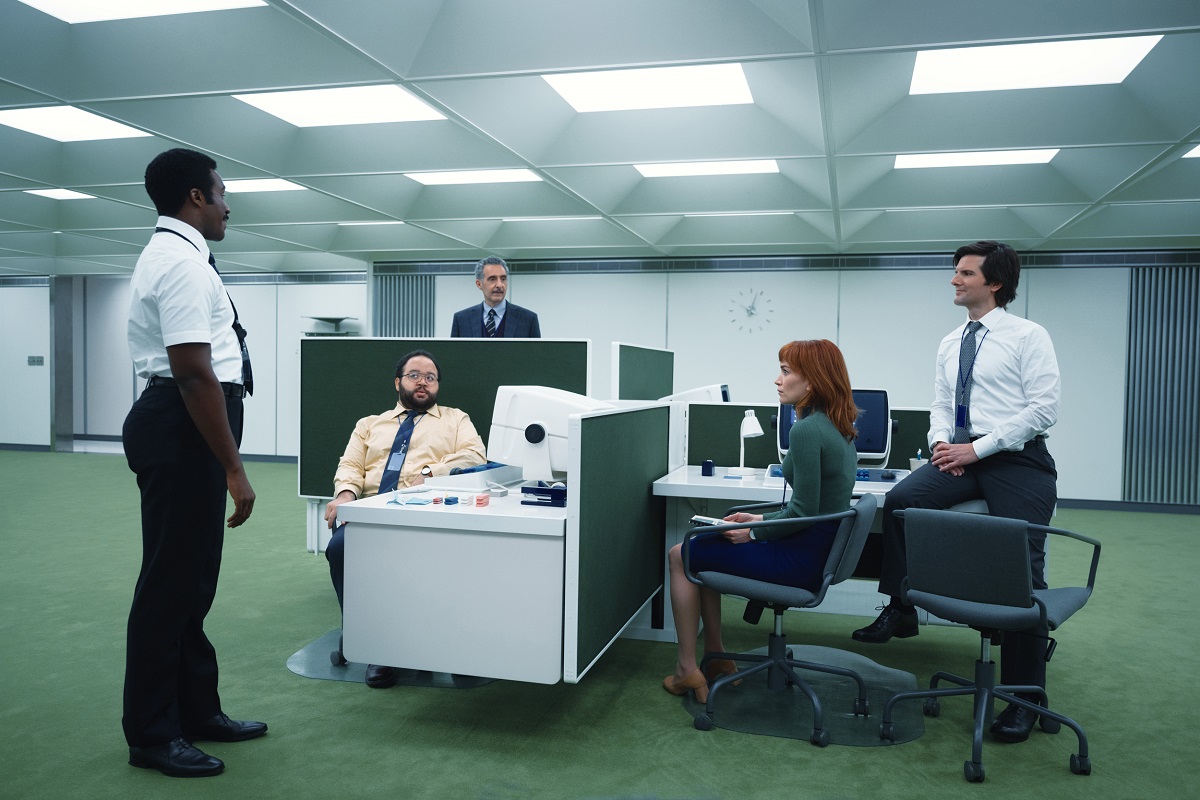 Production still from the show 'Severance' on Apple TV+. In a large, white office with a green carpet and one four-person cubicle station, the four main characters (three men, one woman) are gathered around the cubicles as a Black, male supervisor comes to talk to them.