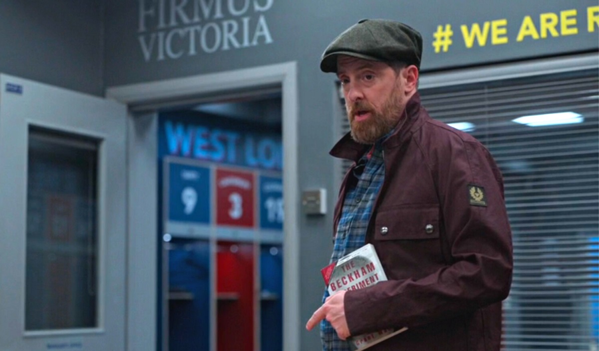 Image of Brendan Hunt as Coach Beard on Apple TV+'s 'Ted Lasso.' He is a white man with a red beard wearing a flat cap hat, a red jacket, and a blue plaid buttondown. He's holding a book: 'The Beckham Experiment' by Grant Wahl as he stands in his office in the Richmond locker room. 