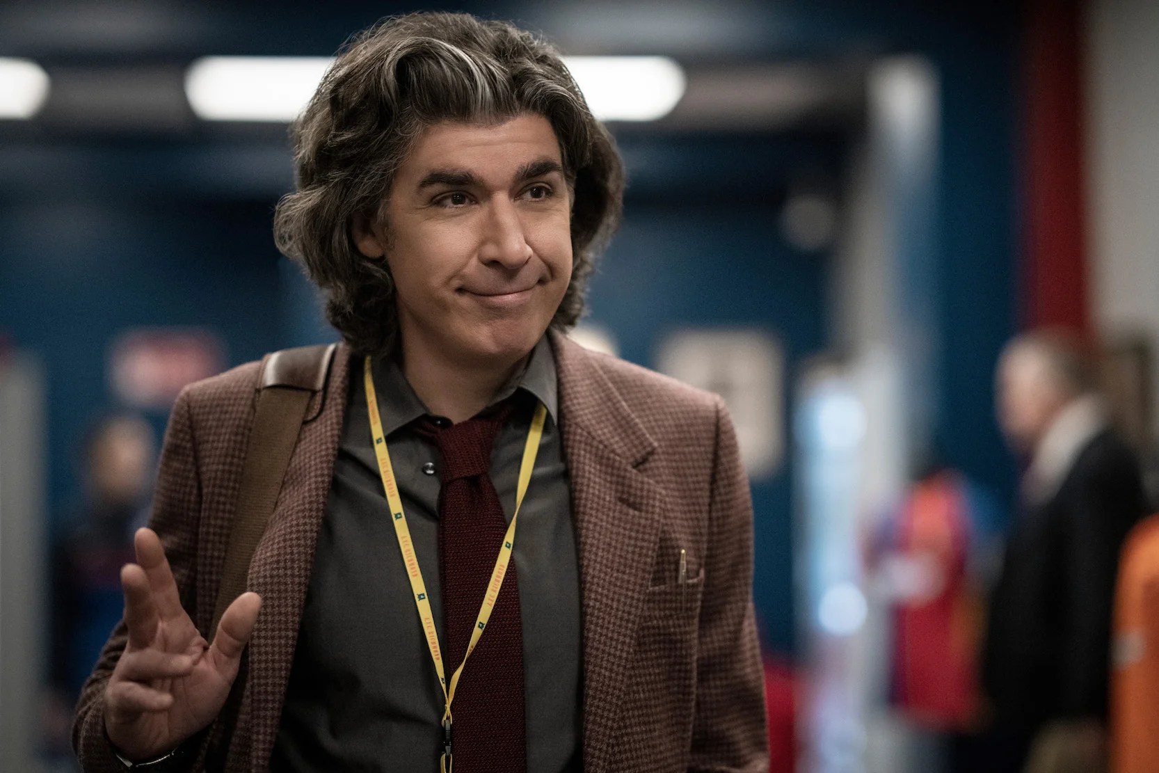 James Lance as journalist Trent Crimm on Apple TV+'s 'Ted Lasso.' He is a white man with thick, chin-length wavy brown hair. He's walking down a hallway in the locker room area of a sports arena wearing a brown, tweed blazer, a green buttondown, and a maroon tie. A yellow lanyard hangs around his neck, and he has a bag hanging from his shoulder by a brown strap. He's waving and giving a closed-mouthed smile to someone. 
