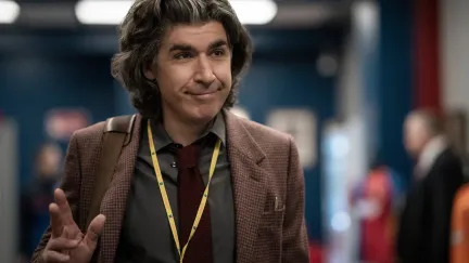 James Lance as journalist Trent Crimm on Apple TV+'s 'Ted Lasso.' He is a white man with thick, chin-length wavy brown hair. He's walking down a hallway in the locker room area of a sports arena wearing a brown, tweed blazer, a green buttondown, and a maroon tie. A yellow lanyard hangs around his neck, and he has a bag hanging from his shoulder by a brown strap. He's waving and giving a closed-mouthed smile to someone.