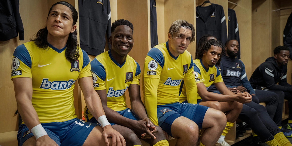 Cristo Fernández as Dani Rojas, Toheeb Jimoh as Sam Obisanya, Phil Dunster as Jamie Tarrt and Moe Hashim as Moe Bumbercatch sitting on a bench in a lockeroom, wearing their yellow and blue away game kits