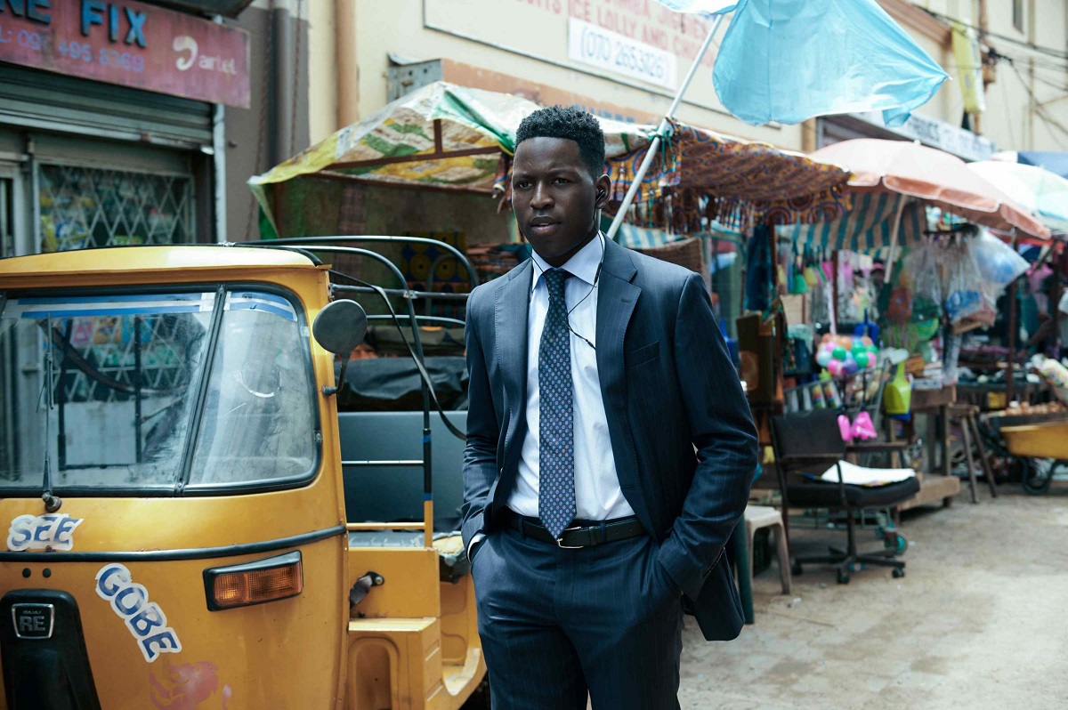 Toheeb Jimoh as Tunde Ojo in Amazon's 'The Power.' He is a Black man wearing a dark blue, pin-striped suit, white button-down, and dark blue tie with white dots on it. He has short hair. He's walking down a street in his city in Nigeria and there are market stalls and a tuk tuk in the background.