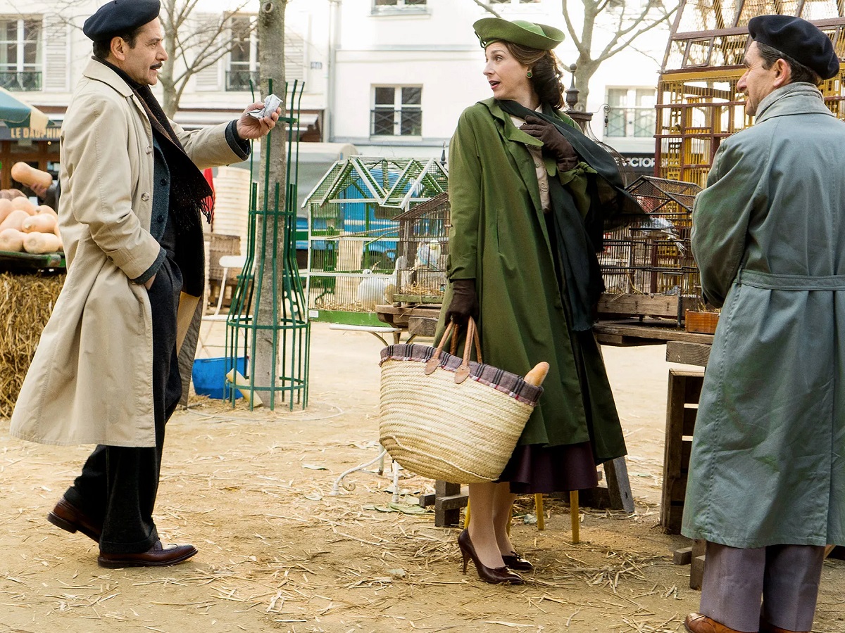 Tony Shaloub as Abe and Marin Hinkle as Rose in a scene from Amazon's 'The Marvelous Mrs. Maisel.' They are in Paris, and Abe, wearing a beret along with his trench coat and dark suit, follows after Rose, who's wearing a green hat and coat with brown gloves and shoes and carrying a wicker tote bag with a baguette in it through a farmer's market.