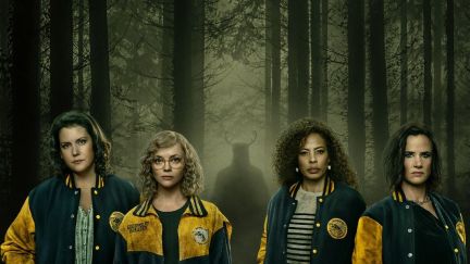 The adult women of 'Yellowjackets' wearing their team jackets and standing in front of a hazy forest with a shadowy figure lurking in the trees