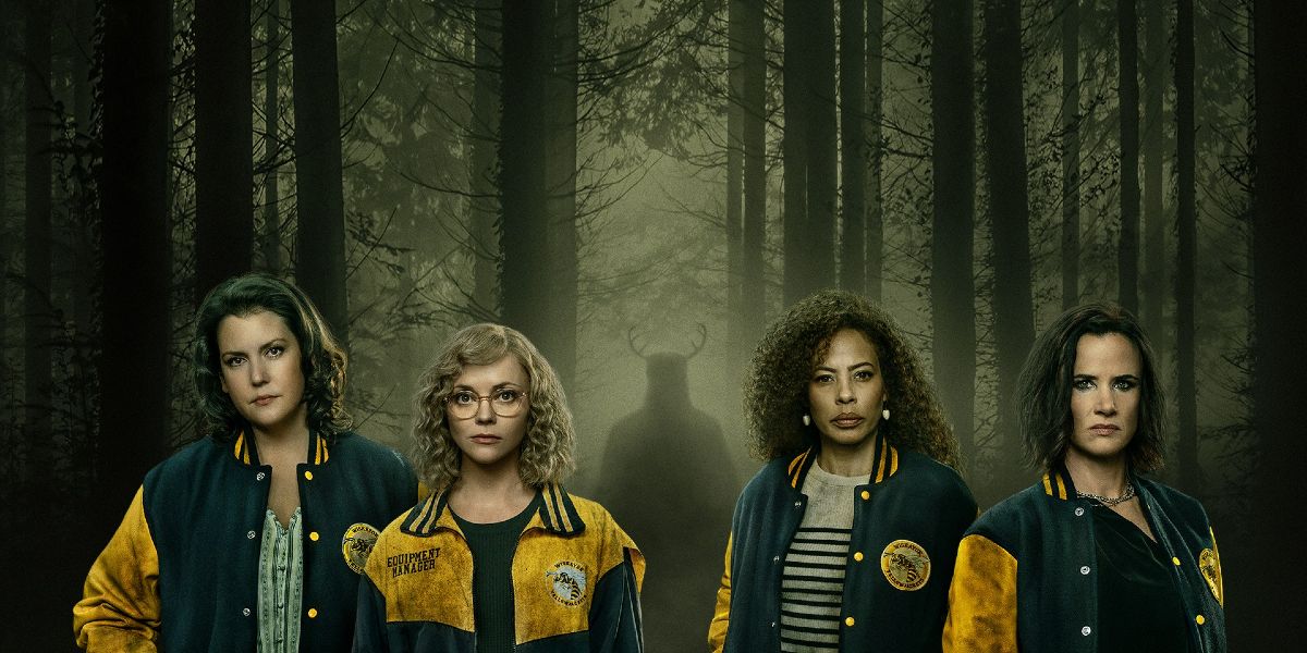 The adult women of 'Yellowjackets' wearing their team jackets and standing in front of a hazy forest with a shadowy figure lurking in the trees