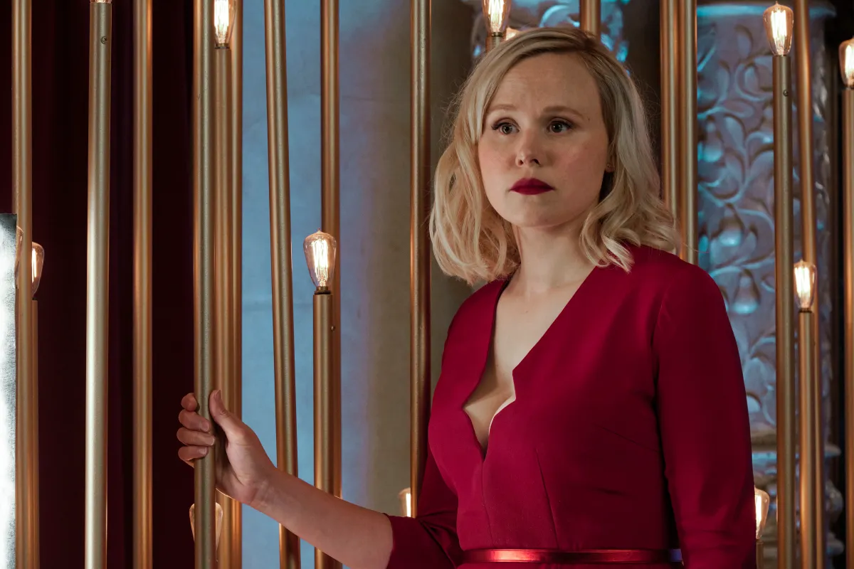 Alison Pill as Agnes Jurati in 'Star Trek: Picard' on Paramount+. She is a white woman with shoulder-length blonde hair wearing a red dress with long sleeves and a low neckline and red lipstick. She's holding onto a bronze light fixture as she looks out at something with a serious expression.