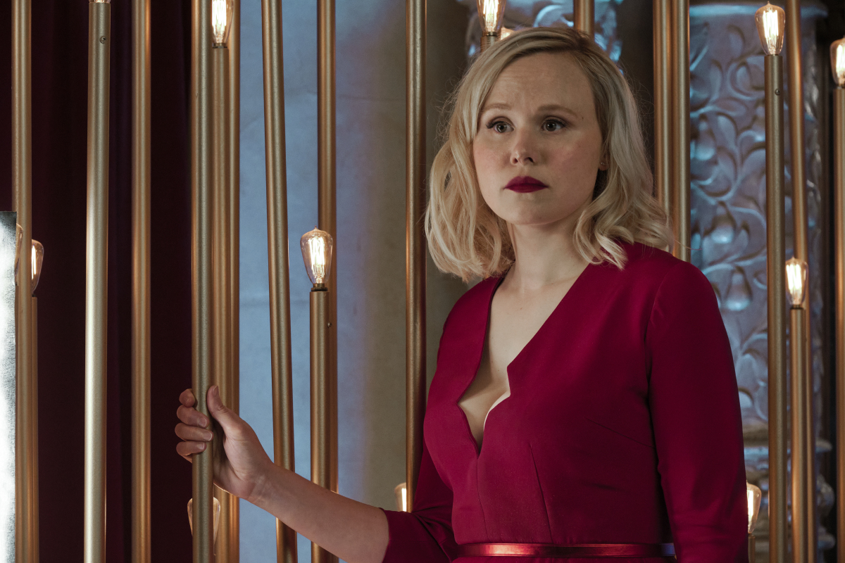 Alison Pill as Agnes Jurati in 'Star Trek: Picard' on Paramount+. She is a white woman with shoulder-length blonde hair wearing a red dress with long sleeves and a low neckline and red lipstick. She's holding onto a bronze light fixture as she looks out at something with a serious expression. 