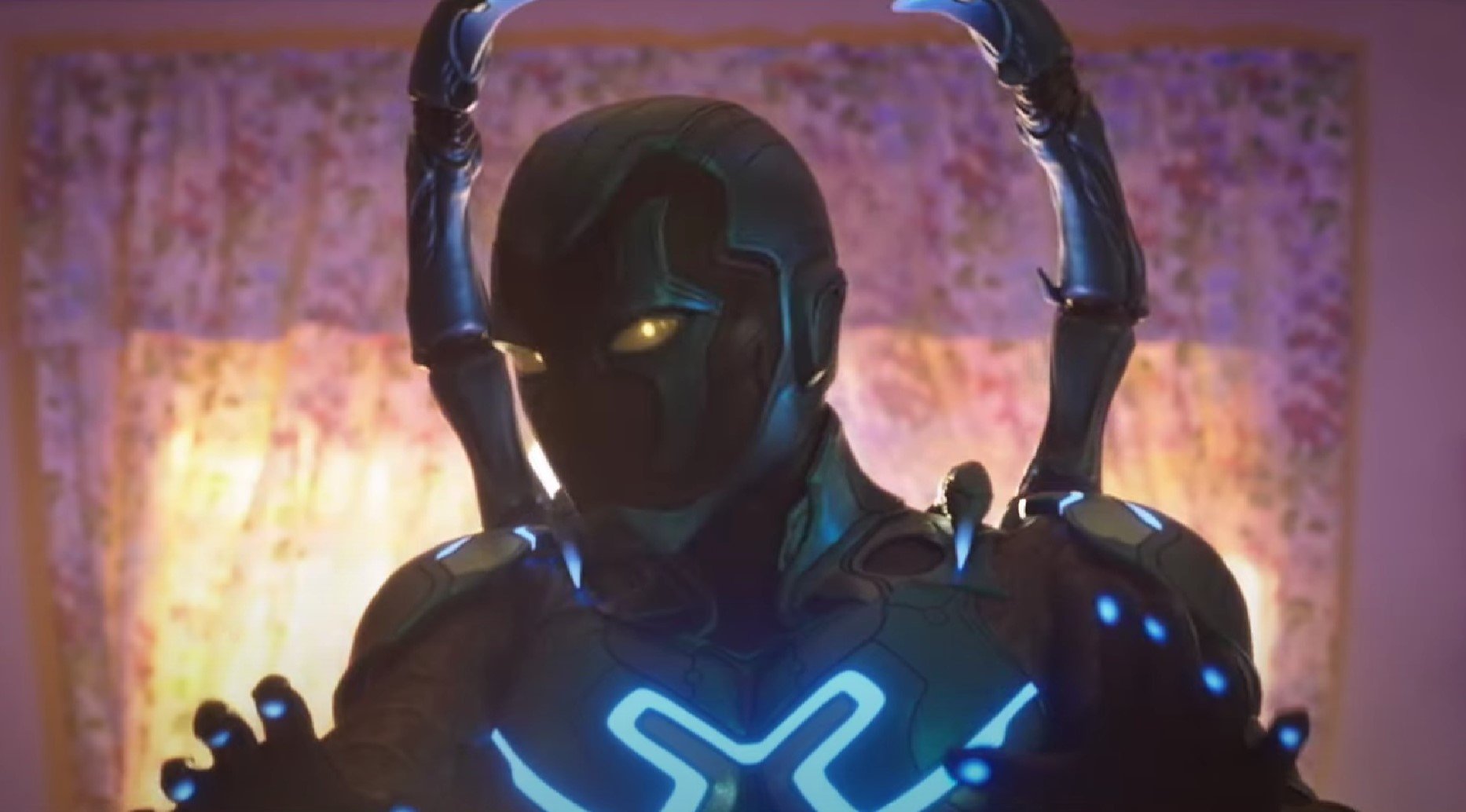 ‎Screenshot of Xolo Maridueña as Blue Beetle in the DC film, 'Blue Beetle.' The suit is blue and black, with parts that glow lighter blue. It has gold eyes, and there are wings coming out of the back. Blue Beetle is holding his hands up reassuringly as he stands in a living room in front of floral curtains. 