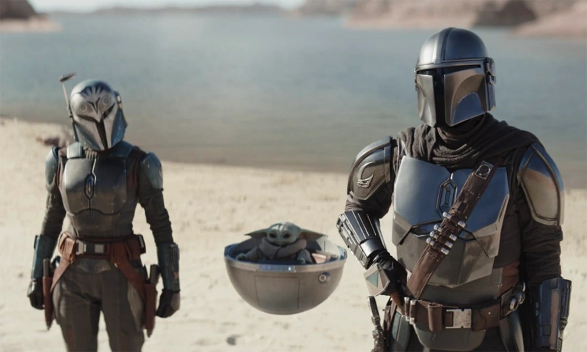 Katee Sackhoff as Bo-Katan, Grogu in his pod, and Pedro Pascal as Din in 'The Mandalorian' on Disney+. They are standing on a beach. Bo-Katan and Din are in their full Mandalorian armor. 
