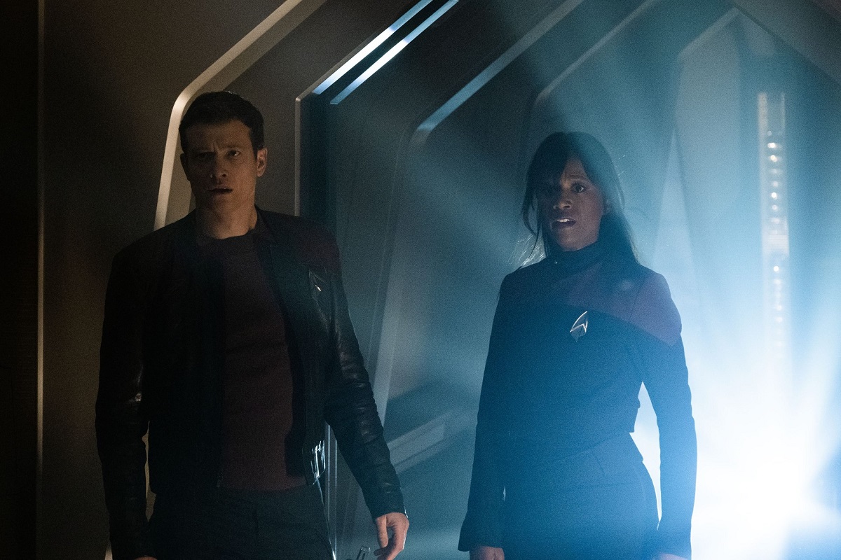 Ed Speleer as Jack Crusher and Ashlei Sharpe Chestnut as Sydney LaForge in a scene from 'Star Trek: Picard" on Paramount Plus. They are walking down a dark ship corridor while back lit by a bright white light. Crusher is a white man with short, dark hair wearing a black, leather jacket, a brown shirt, and black pants. LaForge is a Black woman with long, straight, dark hair with bangs, and she wears a red Starfleet uniform with an Ensign pip on the collar. They're both looking down the corridor in fear. 