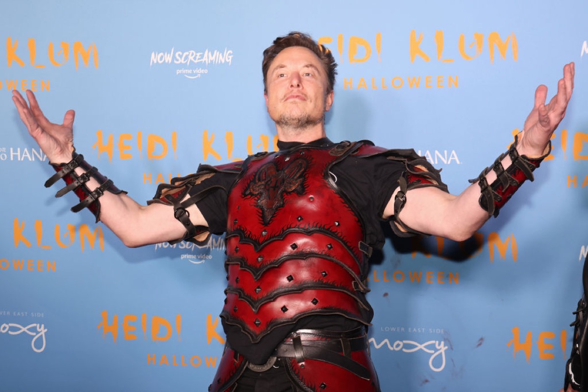 Elon Musk attends Heidi Klum's 2022 Hallowe'en Party. He's wearing some kind of red armor and holding his hands up and puffing his chest out in a smarmy way.