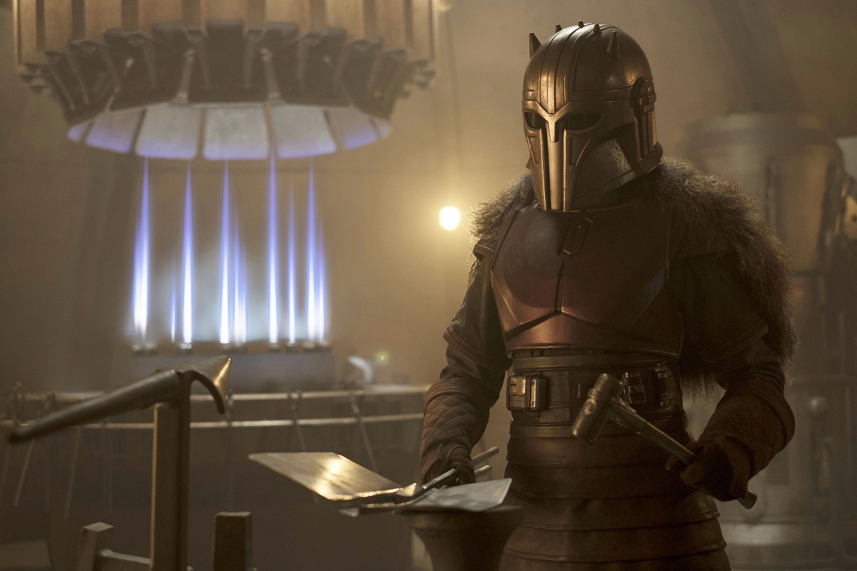 Emily Swallow as The Armorer in a scene from 'The Mandalorian' on Disney+. She is in her full Mandalorian armor, and is holding a hammer, sheet metal, and tongs as she stands in the Forge. 